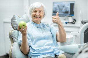 an older woman sitting in the dentist’s chair holding an apple and giving a thumbs up while smiling